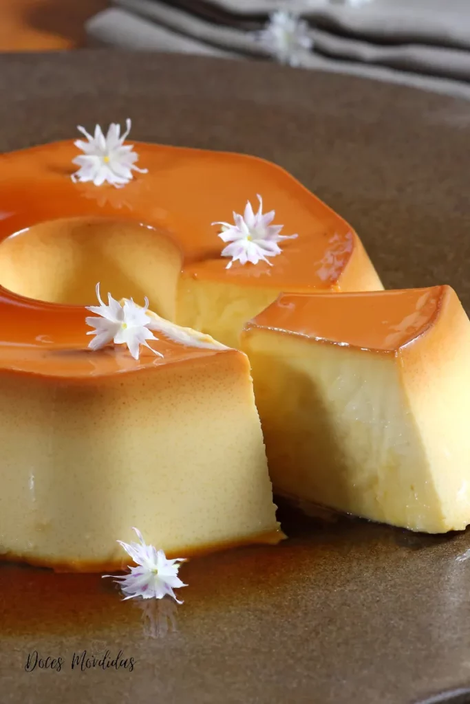 Pudim de leite condensado com calda de caramelo brilhante, decorado com pequenas flores brancas, servido sobre prato marrom — receita do site Doces Mordidas.